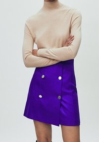 Dopasowany beżowy sweter z golfem, zestawiony z fioletową spódnicą w stylu kopertowym z podwójnymi srebrnymi guzikami, zaprezentowany na modelce z założonymi na siebie ramionami.