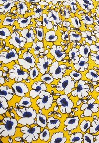 Tissu jaune avec des fleurs blanches bordées de noir et des centres bleus, montrant une couture froncée près du bord supérieur.