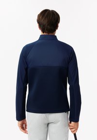 Giacca blu navy realizzata in un tessuto morbido e traspirante. Presenta un colletto alto e maniche aderenti. Il design sul retro include una transizione senza cuciture attraverso le spalle.