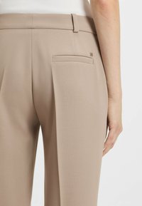 Beige op maat gemaakte broeken met een gladde textuur, voorzien van een achterzak en een klein logo-detail nabij de tailleband.