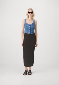 Avklippt denimtop med knäppning, i kombination med en figurnära svart maxikjol. Modellen bär svarta slip-on sandaler och solglasögon.