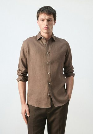 Hombre con camisa marrón de manga larga abotonada y las mangas remangadas, y pantalones marrón oscuro, de pie contra un fondo claro y liso.