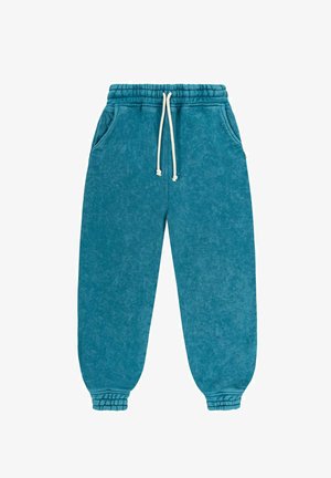 Woman in white top, blue sweatpants, and pink cap leaning against white wall, barefoot next to surfboard on tiled pavement.