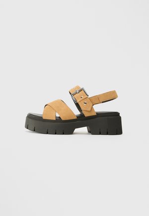 Tan suede sandals with an adjustable buckle, featuring crisscross straps and a chunky black platform sole with a textured finish.