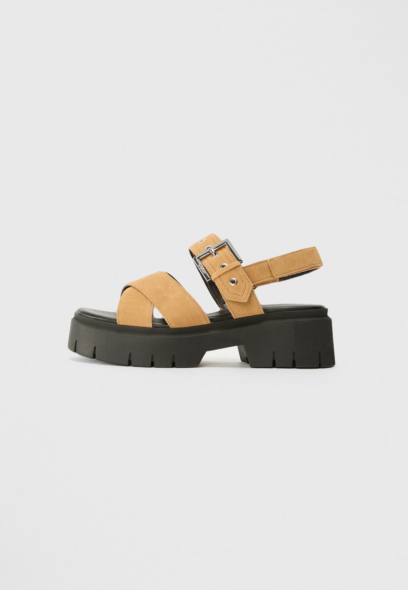 Tan suede sandals with an adjustable buckle, featuring crisscross straps and a chunky black platform sole with a textured finish.