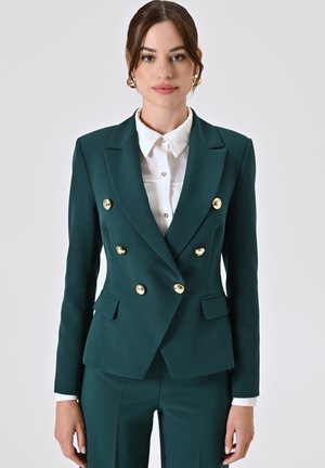 Blazer doppiopetto teal con bottoni dorati, rever a punta e due tasche frontali. Abbinato a una camicia bianca con bottoni sotto.