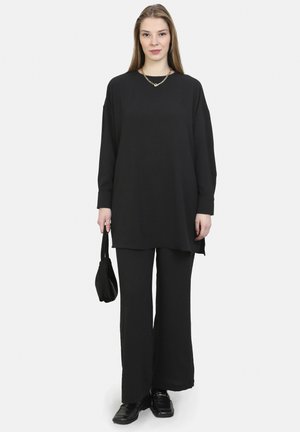 Black oversized long-sleeve top paired with wide-leg pants. Smooth texture, minimalistic design. Accessorized with a small black handbag.