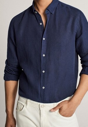Homme portant une chemise bleu marine à manches longues avec les manches retroussées, et un pantalon beige clair avec une main dans la poche.