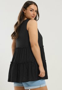 Black sleeveless tunic with tiered ruffles, featuring a textured fabric. The model is wearing blue denim shorts, showcasing the tunic's length.