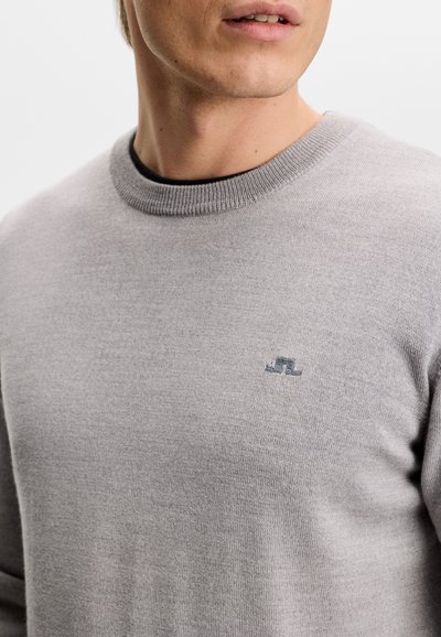 Κοντινό πλάνο ενός ατόμου που φοράει ανοιχτό γκρι πλεκτό πουλόβερ με ένα μικρό κεντητό σκούρο γκρι λογότυπο στο στήθος.