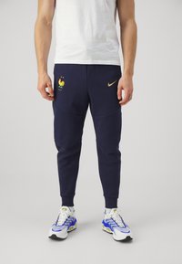 Námořnické jogger kalhoty vyrobené z měkké tkaniny, s zlatým logem Nike a barevným emblemem kohouta. Padnoucí střih s žebrovanými manžetami.