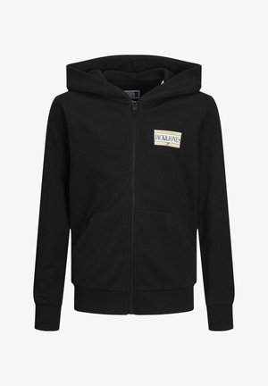 Haut de sweat à zip noir avec poches avant et patch logo, pantalon cargo beige avec poches latérales, et baskets noires avec rayures blanches.