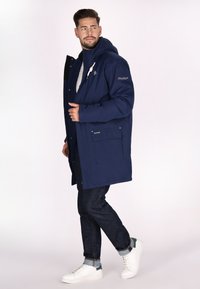 Parka impermeabile blu navy con cappuccio, chiusura con zip, tasca frontale e fodera nera. Abbinata a jeans scuri e sneakers bianche.