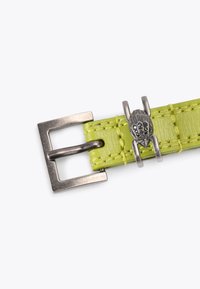 Zielony skórzany pasek o plecionym wzorze, srebrna metalowa klamra i dekoracyjny charms w kształcie dłoni, z czarnymi detalami.