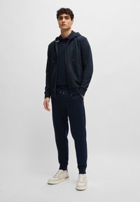 Fleece azul marinho com cordões, combinado com uma polo correspondente e calças de jogging. Apresenta punhos elásticos, design minimalista e ténis em tons neutros.