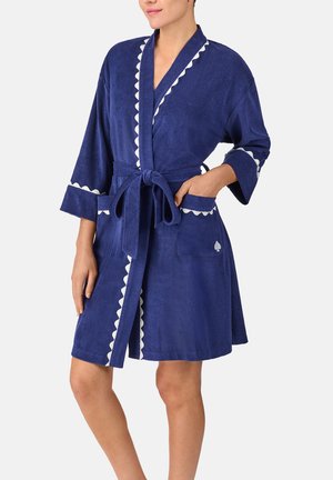 Mujer vestida con una bata azul marino hasta la rodilla, con bordes blancos en forma de concha, atada a la cintura, con las manos en los bolsillos, de pie frente a un fondo liso.