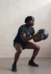 Svart atletisk skjorte og shorts, crouches med en svart medisinball i et innendørs miljø, teksturert gulv, lys vegg som bakgrunn.
