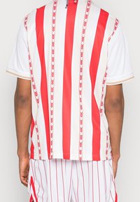  camiseta de manga corta con rayas verticales rojas y blancas, con una tela texturizada y acentos en patrón de flechas, que exhibe un diseño deportivo.