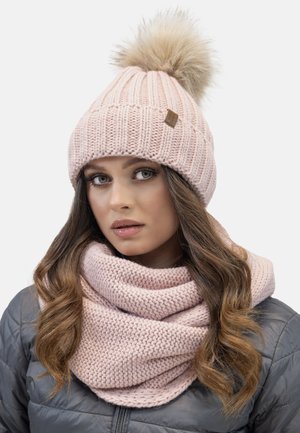 Cappello lavorato a maglia di un delicato rosa chiaro con un pompon in pelliccia sintetica e un'etichetta in pelle. Abbinato a una sciarpa lavorata a maglia spessa dello stesso rosa chiaro.