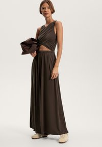 Braunes Maxikleid mit einem verdrehten Frontdesign, ärmelloser Schnitt und elastischem Bund. Begleitet von einer passenden braunen Jacke.