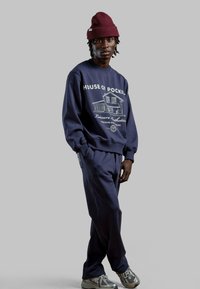 Navy blauwe sweatshirt met "HOUSE OF POCKIL" graphic en bijpassende broek, gecombineerd met een kastanjebruine beanie en zilveren sneakers. Zacht materiaal, casual pasvorm.