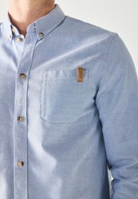 Chemise bleu clair boutonnée avec poche poitrine et boutons marron portée par une personne, montrant le haut du torse et le décolleté.