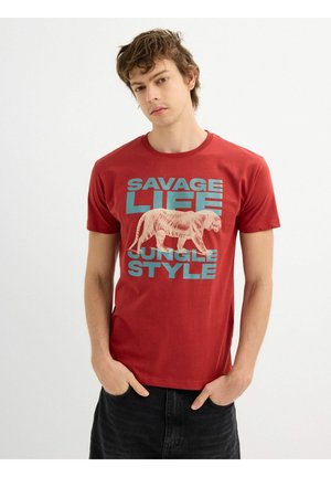 Camiseta de algodón roja con un gráfico de tigre en color crema y un texto en negrita color teal que dice "SAVAGE LIFE JUNGLE STYLE". Manga corta y cuello redondo.