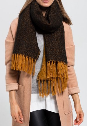 Femme portant une grande écharpe noire et moutarde à franges, un manteau rose clair, un pull gris, une chemise blanche et un pantalon noir.