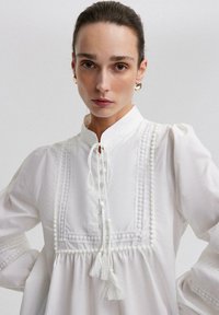 Camicia bianca in cotone con colletto alto, rifiniture decorative, maniche a sbuffo e dettagli in frangia sul collo e sull'orlo.