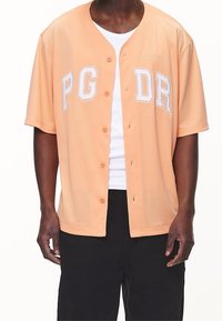 Camicia a maniche corte color pesca con bottoni, caratterizzata dalla scritta bianca "PG DR", vestibilità rilassata e texture a rete. Indossata sopra una canotta bianca.