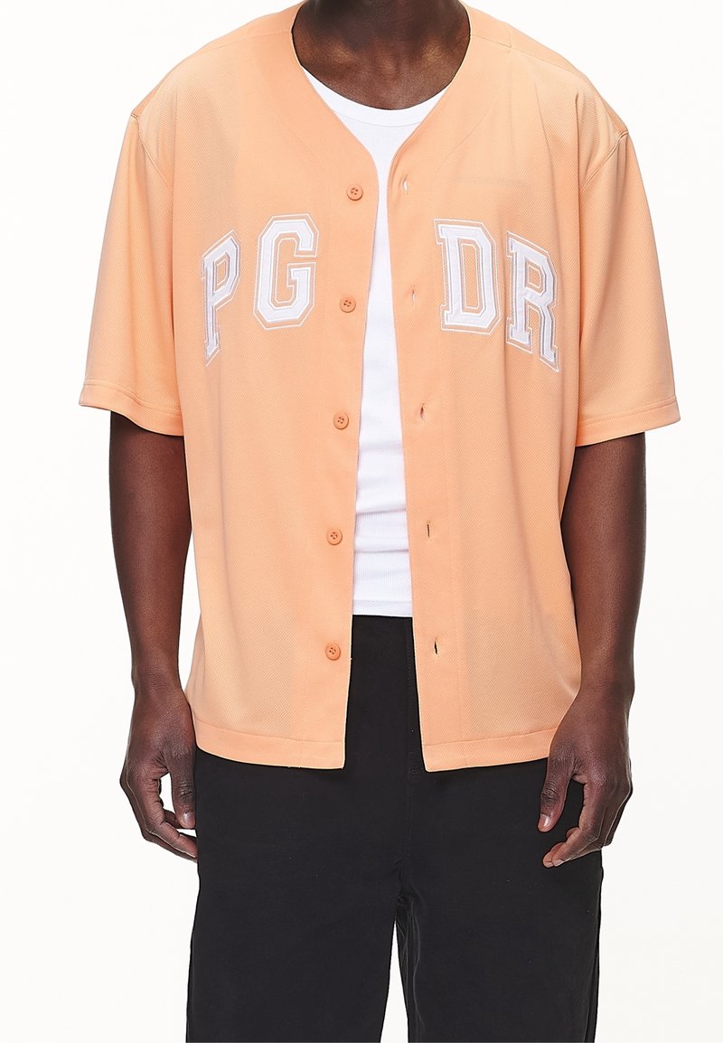 Camicia a maniche corte color pesca con bottoni, caratterizzata dalla scritta bianca "PG DR", vestibilità rilassata e texture a rete. Indossata sopra una canotta bianca.