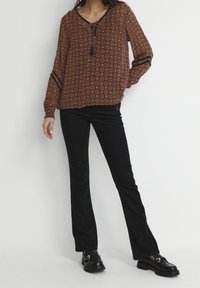 Blusa marrone a fantasia con maniche lunghe, collo con stringa e dettagli neri, abbinata a pantaloni neri a zampa d'elefante e mocassini neri spessi.