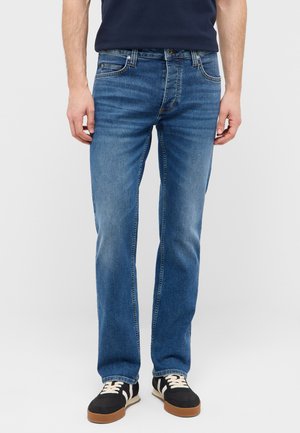 Mann trägt mittelblaue Jeans mit geradem Bein, schwarze Sneakers mit weißen Schnürsenkeln und ein dunkelblaues Hemd, steht vor einem schlichten hellen Hintergrund.