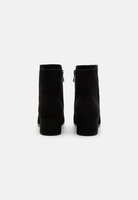 Botas de ante negras hasta el tobillo con un tacón cuadrado, que cuentan con un cierre de cremallera lateral y una textura suave. El diseño es minimalista y elegante.