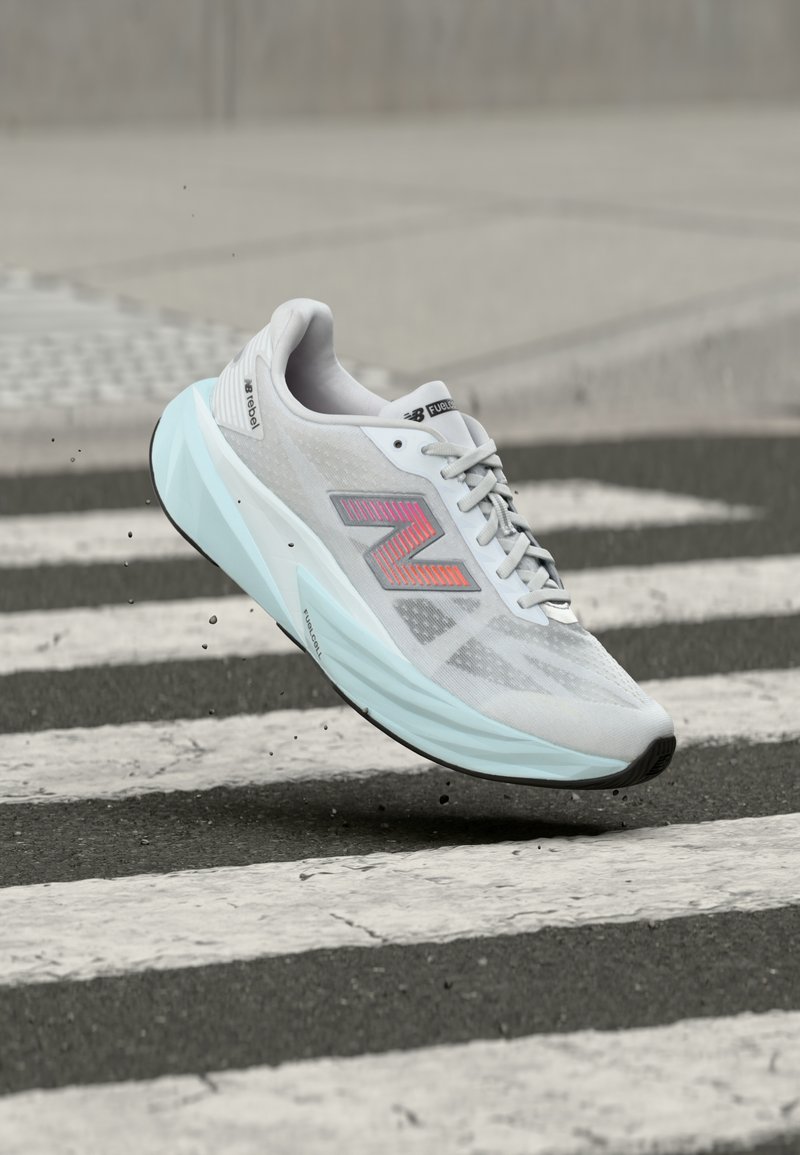 Λευκό και γαλάζιο αθλητικό παπούτσι New Balance με ροζ λογότυπο, στον αέρα πάνω από διάβαση πεζών σε ασφαλτοστρωμένο δρόμο.