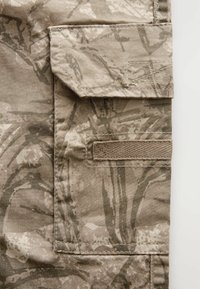 Tessuto camouflage color sabbia con tasca a toppa e cuciture testurizzate. Il design presenta tonalità variabili e motivi astratti.