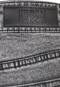 Sivi traper tkanina s izraženom teksturom, s crnom kožnom etiketom utisnutom s tekstom "URBAN CLASSICS DENIM FACTORY" i kontrastnim šavovima.