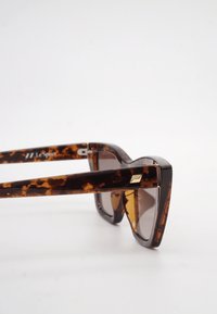 Σκουρόχρωμα γυαλιά ηλίου με μοτίβο χελώνας σε γωνιώδη πλαίσια και μικρό χρυσό ορθογώνιο διακοσμητικό στο μπράτσο, εμφανιζόμενα σε λευκό φόντο.