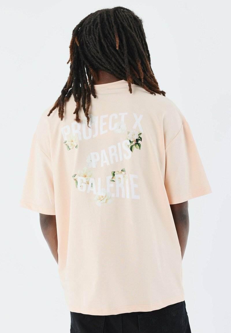 Perzikkleurig oversized t-shirt met witte tekst "PROJECT X PARIS GALERIE" en bloemaccenten. Katoenen materiaal, korte mouwen, ontspannen pasvorm.