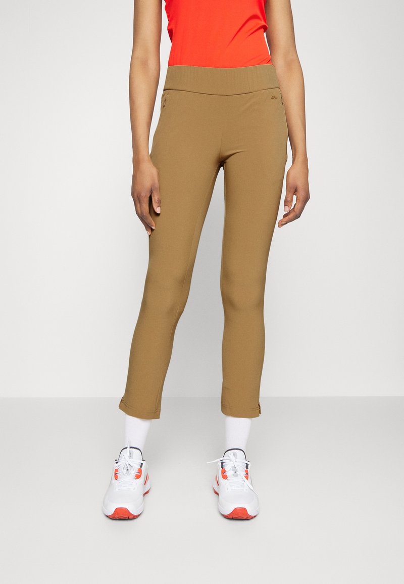 Pantalones marrones ajustados hasta el tobillo con cintura elástica, con bolsillos laterales. Combinados con zapatillas deportivas blancas y naranjas.