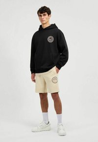Sudadera negra con capucha y bolsillo frontal, combinada con pantalones cortos beige con un logo circular. El modelo lleva zapatillas blancas y calcetines altos.