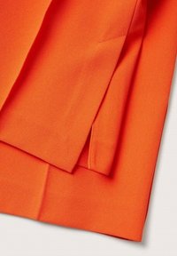 Tissu orange vif avec des plis nets et précis ainsi que des détails de couture, montrant la texture et des bords ajustés sur une surface lisse.