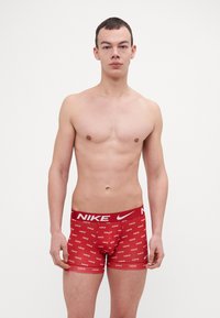 Czerwone majtki bokserki z markowym paskiem. Materiał wydaje się gładki. Posiadają powtarzający się nadruk białego logo Nike na całej tkaninie.