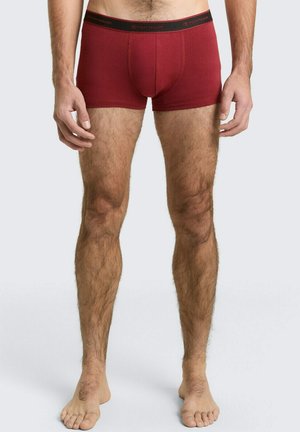 Mann trägt rote Boxershorts mit schwarzem Bund mit der Aufschrift „Tom Tailor“, steht barfuß vor einem einfarbigen hellen Hintergrund.
