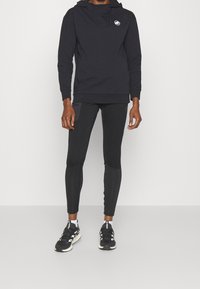 Sudadera con capucha negra con un pequeño logo, combinada con leggings negros que presentan paneles texturizados y zapatillas deportivas negras con detalles blancos.