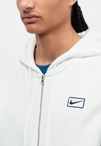 Biela zipsova mikina so strieborným zipsom, s námořníčko-modrou vyšitou logom Nike na ľavej hrudi a modrou košeľou pod ňou.