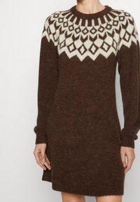 Robe tricotée marron avec un empiècement crème à motifs. Elle possède des manches longues et un col côtelé. Texture douce, coupe décontractée, design longueur genou.