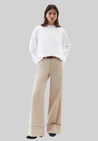 Bílá mikina s dlouhým rukávem v kombinaci s béžovými kalhotami s vysokým pasem a širokými nohavicemi, zakončenými ohrnutým lemem a hnědým páskem, na šedém pozadí.