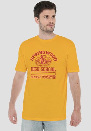 Mann trägt gelbes T-Shirt mit rotem Schriftzug "Springwood High School Physical Education" und Schlangen-Emblem, steht vor neutralem Hintergrund.