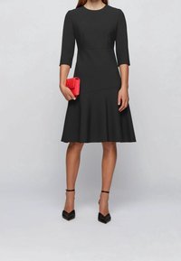 Svart klänning med trekvartslånga ärmar, figurnära överdel, utsvängd kjol och slätt tyg. Ett par svarta klackskor och en röd clutchtväsk.